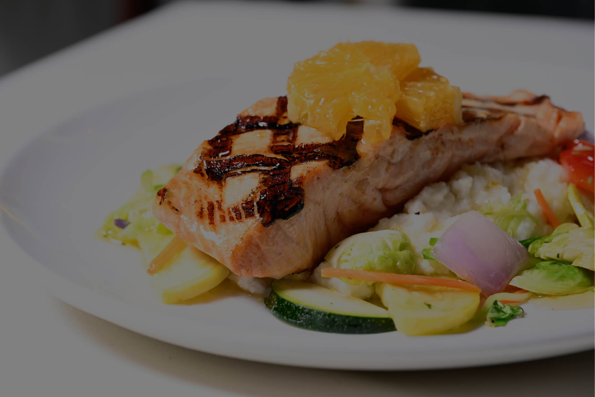 Image of cooked salmon on plate with oranges on top of it while sitting on a bed of cooked vegetables